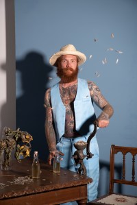 ein tättowierterrotbärtiger Mann steht mit einem blauen Anzug an einer Getreidemühle neben ihm vertrocknete Sonnenblumen, Sonnenblumenkerne fliegen durch die Luft.